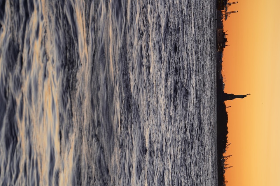 The Statue of Liberty, at dusk, in the distance, across the water. The statue is visible as a silouhette against the yellow orange sky.